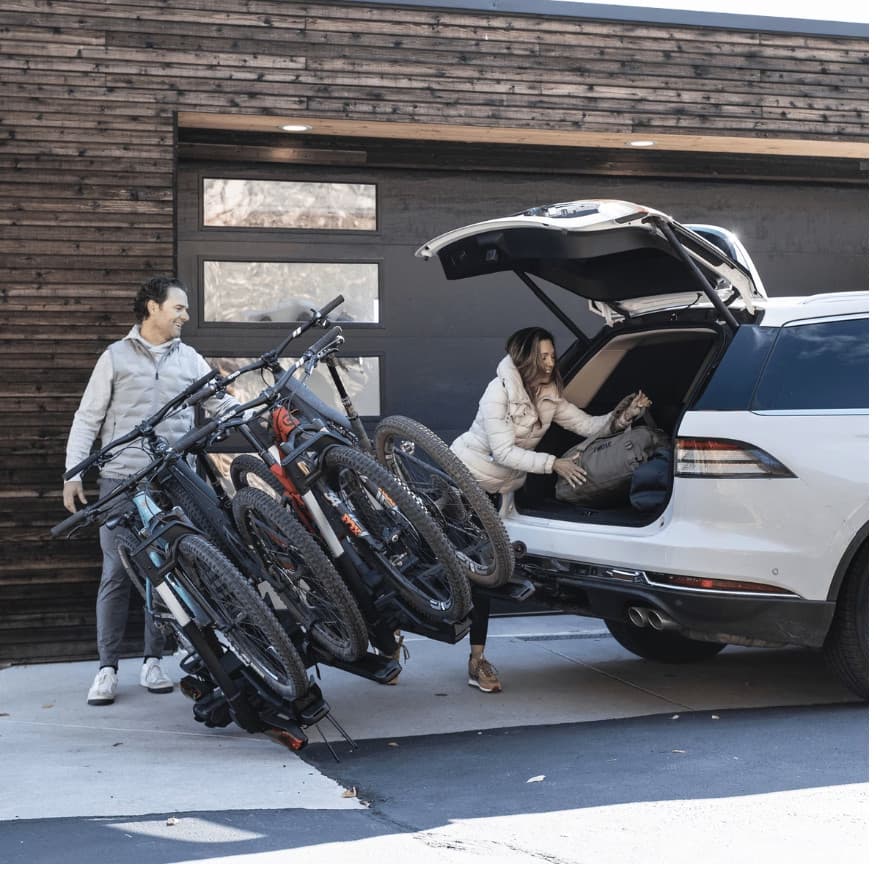 Thule Verse Add-On fully loaded with 4 bikes installed on SUV. Verse is tilted down to show trunk access for woman rummaging through trunk of SUV